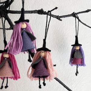 May include: Four hanging ornaments shaped like figures wearing black pointed hats. Each figure has a wooden bead head, yarn hair in shades of pink and purple, and a felt dress in varying tones of pink, purple, and grey with thin black string legs.