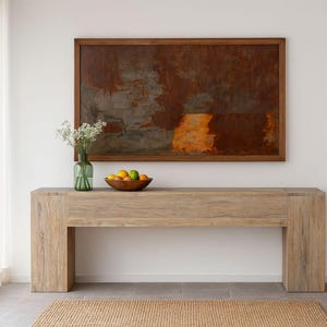 Könnte beinhalten: Eine hellfarbene Holzkonsolentisch mit rechteckiger Platte und Blockbeinen. Darüber ein gerahmtes abstraktes Gemälde in Braun- und Orangetönen. Eine grüne Glasvase mit weißen Blumen und eine Holzschale mit Obst stehen auf dem Tisch.