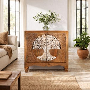 Op de afbeelding: Een bruine houten kast met een uitgesneden boomontwerp op de deuren. De boomsculptuur is wit. Een vaas met groene takken staat erop. De kast staat in een kamer met een bank en een plant in een pot.
