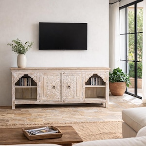 Puede incluir: Un mueble de televisión largo de madera clara con dos armarios centrales con intrincados patrones florales tallados y dos estantes laterales abiertos. El mueble tiene una superficie rectangular y se apoya sobre patas cortas.