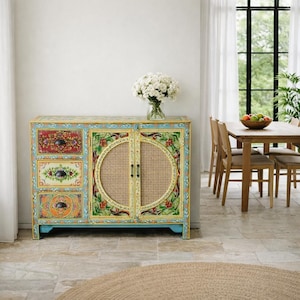 Może przedstawiać: Ręcznie malowana szafka z trzema szufladami i dwiema drzwiami. Szafka posiada kwiatowy wzór w kolorze czerwonym, zielonym i żółtym, z plecionym rattanem pośrodku drzwi. Szafka stoi na niebieskiej podstawie.