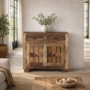 Könnte beinhalten: Ein rustikaler Holzschrank mit zwei Schubladen und zwei Türen. Der Schrank besteht aus Altholz mit sichtbaren Knoten und Maserungen. Die Türen haben Metallringgriffe und dekorative Holzpaneele. Oben steht eine Vase mit Grün.