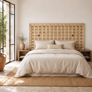 May include: A large rectangular wooden headboard featuring a grid of carved square panels with floral motifs. The bed is dressed in light beige linen bedding, including two large pillows and a smaller rectangular bolster pillow in the center.