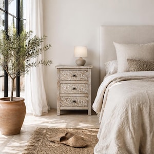 Puede incluir: Una mesita de noche de madera de tres cajones con intrincados patrones florales tallados en los frentes, con una pequeña lámpara de mesa con pantalla blanca encima, junto a una cama con ropa de cama beige y un par de zapatillas tejidas sobre una alfombra.