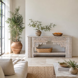 Peut inclure: Une console en bois clair avec des détails sculptés complexes sur les pieds et le tablier, dotée d'une étagère inférieure contenant un panier tressé. La table présente un plateau rectangulaire et une finition patinée sur un sol clair.