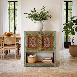 May include: A rectangular wooden cabinet featuring two doors with intricate hand-painted floral and geometric patterns in red, green, and blue. The cabinet includes an open lower shelf containing a woven basket and a stack of books.