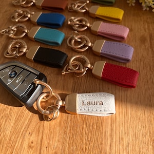 May include: A collection of keychains with different coloured leather straps and gold metal rings. The straps are decorated with stitching and some have a name engraved on them. The keychains are arranged on a wooden surface.