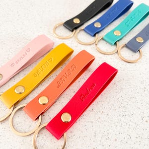 Puede incluir: Un conjunto de llaveros de cuero de colores con nombres personalizados y un símbolo de corazón. Los llaveros están unidos a llaveros de oro.
