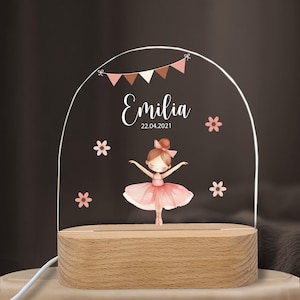 Może przedstawiać: Dekoracyjna lampka nocna z motywem baletnicy. Akrylowy panel wyświetla imię "Emilia" i datę "22.04.2021", z kwiatowymi akcentami i banerem. Światło stoi na drewnianej podstawie.