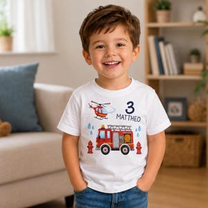 Puede incluir: Un niño pequeño lleva una camiseta blanca con un diseño colorido de un camión de bomberos, un helicóptero y una boca de incendios. El número 3 y el nombre "MATTHEO" están impresos en la camiseta. El niño sonríe.