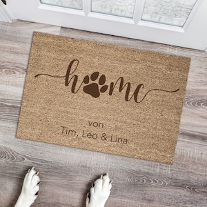 Könnte beinhalten: Eine rechteckige Fußmatte mit dem Wort "home" in Schreibschrift und einem Pfotenabdruck-Design. Der Text "von Tim, Leo & Lina" steht darunter. Die Matte ist naturfarben und liegt auf einem Holzboden.
