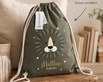 May include: An olive green drawstring backpack featuring a stylized lion face graphic, the name "Mattheo," and the date "23.08.2021" printed in white and gold. The bag has thick cream-colored rope straps and an EarthAware brand tag attached.
