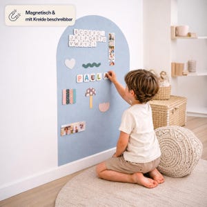 Op de afbeelding: Een boogvormige, lichtblauwe magnetische muursticker met een matte afwerking. Het oppervlak bevat diverse magnetische vormen, waaronder alfabetletters, een paddenstoel, een hart en de naam PAULA. Een tekstvakje bovenaan toont Magnetisch & mit Kreide beschreibbar.