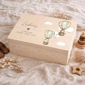 May include: A light-colored wooden box with a hinged lid. The lid is decorated with two hot air balloon illustrations and the name "Lukas" in script. The box also includes the date, time, weight (3590 g) and height (55 cm) of a baby.