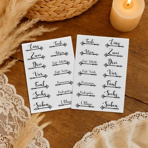 Puede incluir: Dos hojas de pegatinas blancas con texto de caligrafía negra. El texto incluye números y frases como "Taufe", "Sitzen" y "Erste Schritte". Una vela encendida y elementos decorativos están en el fondo.