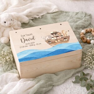 May include: A wooden keepsake box with a painted illustration of Noah's Ark, featuring various animals and blue watercolor waves. The box has the name "David" and other details printed on the lid. The box is made of light-colored wood.