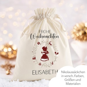 Puede incluir: Una bolsa de regalo crema con cordón. Las palabras "FROHE WEIHNACHTEN" están escritas en negro. Un diseño de corona circular con la silueta de una niña, velas y un reno está impreso debajo. El nombre "ELISABETH" está impreso debajo.