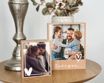 Op de afbeelding: Twee houten fotolijsten met witte harten en de tekst "Ik hou van je" onderaan een lijst. De lijsten staan op een houten tafel en bevatten foto's van families.
