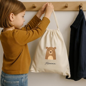 Puede incluir: Una mochila beige con cord&oacute;n y una ilustraci&oacute;n de oso marr&oacute;n con el nombre "Hanna" en cursiva. La bolsa est&aacute; colgada de un perchero de madera. A la derecha cuelga una chaqueta azul marino.