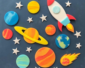 May include: A blue background with a rocket, planets, and stars made of fondant. The planets are orange, yellow, red, and blue with swirls of colour. The rocket is white with red and blue accents. The stars are white.