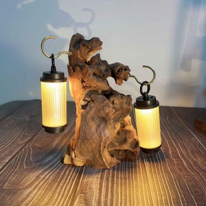 May include: A unique wooden sculpture with two hanging lanterns. The lanterns have a black top and bottom with a ribbed, cylindrical, yellow-tinted glass. The lanterns hang from brass-colored hooks attached to the wood. The wood is a natural, light brown color.