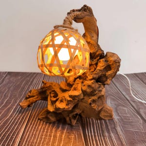 May include: A unique table lamp featuring a woven bamboo shade suspended from a natural wood base. The lamp's shade is a warm orange color, and the base has a rustic, textured appearance. The lamp is plugged in and lit, casting a warm glow.