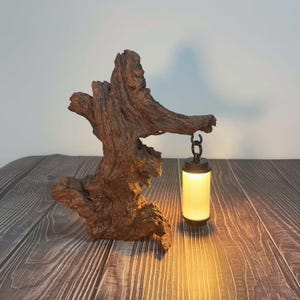May include: A unique lamp featuring a natural wood base with a hanging lantern. The wood has a rough, textured appearance with various shades of brown. The lantern is cylindrical, with a warm, yellow glow emanating from within, creating a cozy ambiance.