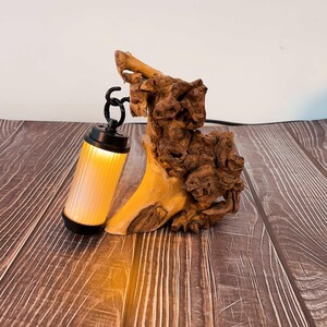May include: A unique table lamp featuring a natural wood base and a cylindrical light fixture. The light has a black metal frame and a ribbed, translucent shade that emits a warm, yellow glow. The wood base has a light brown color.