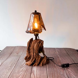 May include: A unique table lamp featuring a brown, textured driftwood base. The lamp has a dark amber-colored glass shade with a wavy edge. The bulb is lit, casting a warm glow. The lamp is connected to a black cord with an on/off switch.
