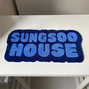 May include: A blue rectangular rug featuring the text "SUNGS00 HOUSE" in a lighter blue, rounded font. The rug has a dark blue border and a tag attached.
