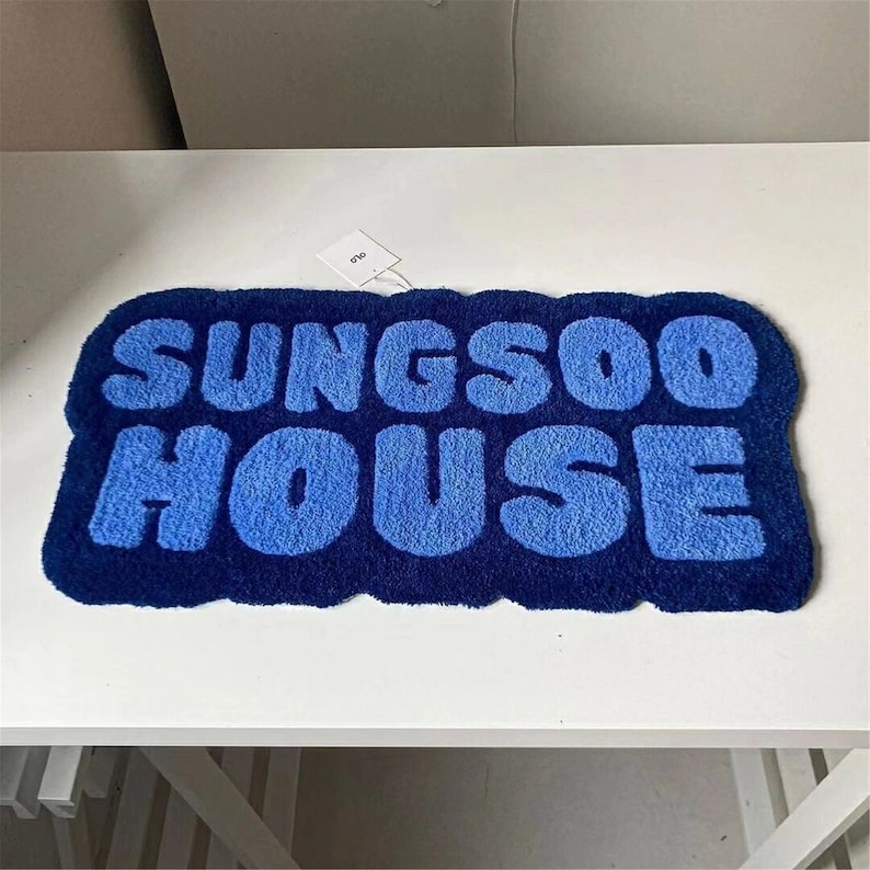 May include: A blue rectangular rug featuring the text "SUNGS00 HOUSE" in a lighter blue, rounded font. The rug has a dark blue border and a tag attached.