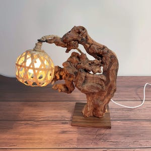 May include: A unique wooden table lamp featuring a woven bamboo shade. The lamp is crafted from a natural, textured piece of wood, mounted on a square wooden base. The shade is suspended by a rope, and the lamp emits a warm, inviting glow.