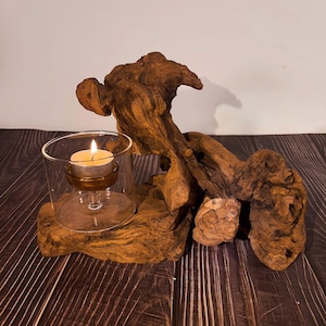 Könnte beinhalten: Ein rustikaler Kerzenhalter mit einer brennenden Teelichtkerze in einem Glasbecher, auf einem Stück Naturholz. Das Holz hat eine satte braune Farbe und eine einzigartige, strukturierte Form. Ein größeres Stück Treibholz befindet sich hinter dem Kerzenhalter.