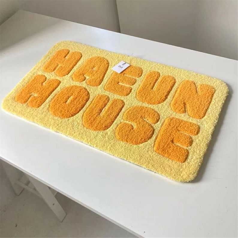 May include: A rectangular yellow rug with the words "HAEUN HOUSE" in orange, fluffy, bubble letters. The rug is placed on a white surface.