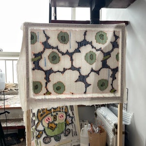 May include: A textile art piece in progress, showing a floral design with white, green, and navy blue colours. The artwork is stretched on a wooden frame. A second, smaller artwork is visible below. The background includes a spiral staircase.