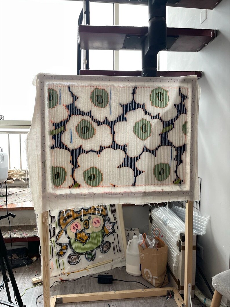 May include: A textile art piece in progress, showing a floral design with white, green, and navy blue colours. The artwork is stretched on a wooden frame. A second, smaller artwork is visible below. The background includes a spiral staircase.