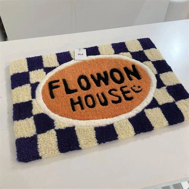 May include: A rectangular, checkered rug with a navy blue and cream pattern. The centre features an orange oval with the words "FLOWON HOUSE" in black, and a smiling face. The rug is made of a soft, textured material.