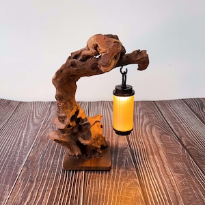 May include: A unique table lamp featuring a natural wood base and a decorative branch supporting a hanging lantern. The lantern has a black top and bottom with a yellow, cylindrical shade. The lamp is a warm, inviting accent piece.
