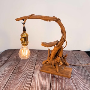 May include: A handcrafted wooden table lamp with a natural driftwood design. The lamp has a curved wooden arm supporting a brass bulb socket and a vintage-style Edison bulb. The base is a rectangular wooden block. The lamp is plugged in.