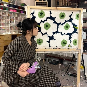 May include: A person working on a large rug with a floral design in white, green, and navy blue, mounted on a wooden frame. They are holding a purple tufting gun. The room has shelves of yarn and craft supplies.