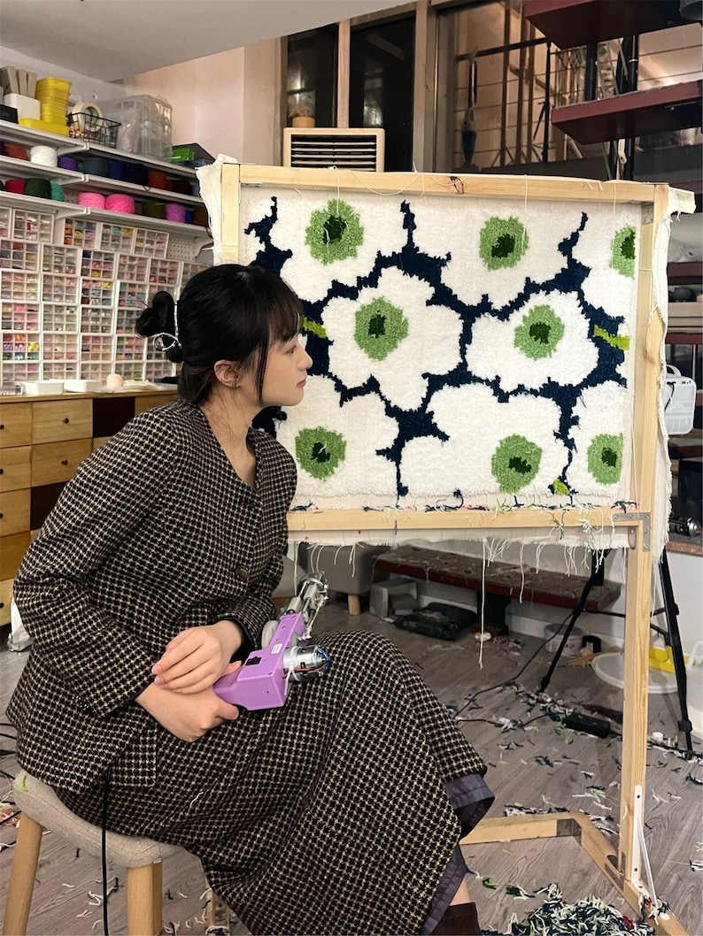 May include: A person working on a large rug with a floral design in white, green, and navy blue, mounted on a wooden frame. They are holding a purple tufting gun. The room has shelves of yarn and craft supplies.