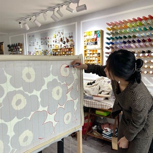 May include: A person working on a large textile art piece with a floral pattern. The design features grey flowers with red outlines on a light background. The artist is using a red marker. The room has spools of thread and other craft supplies.