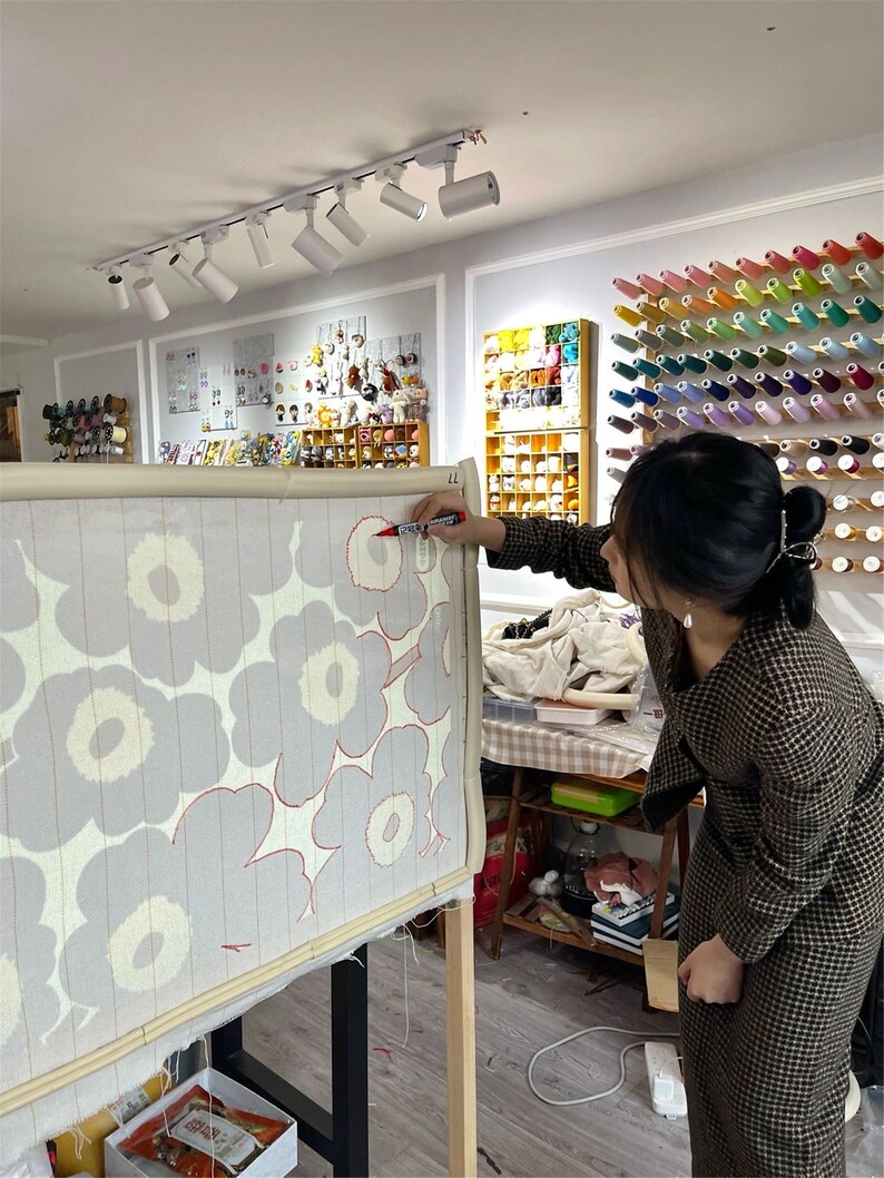 May include: A person working on a large textile art piece with a floral pattern. The design features grey flowers with red outlines on a light background. The artist is using a red marker. The room has spools of thread and other craft supplies.