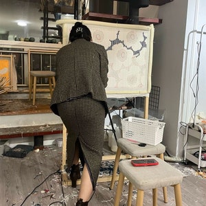 May include: A person working on a large rug with a geometric design. The rug is stretched on a wooden frame. The person wears a brown and black checked suit and brown boots. The room has a modern, artistic vibe, with a spiral staircase.