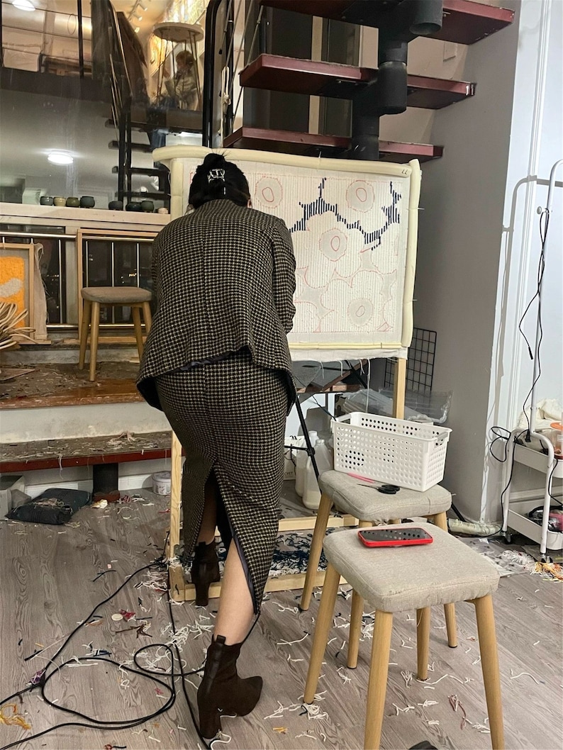 May include: A person working on a large rug with a geometric design. The rug is stretched on a wooden frame. The person wears a brown and black checked suit and brown boots. The room has a modern, artistic vibe, with a spiral staircase.