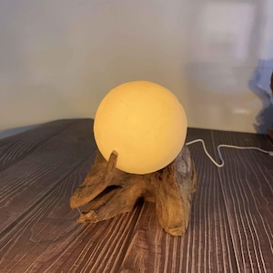 May include: A round, glowing, yellow-toned lamp sits atop a piece of natural driftwood. The lamp has a spherical shape and emits a soft light. The driftwood base has a natural, textured appearance.