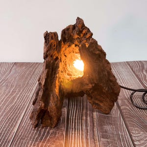 May include: A unique, handcrafted wooden lamp. The lamp is made from a piece of natural wood, with a hollowed-out center that emits a warm, inviting glow. The wood has a rich, brown color and a textured surface. The lamp is plugged in with a black cord.