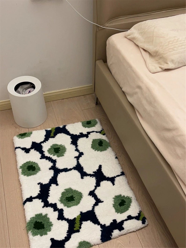 May include: A rectangular rug with a floral design. The rug displays large white flowers with green centres and dark blue outlines. It's placed on a light wooden floor, near a bed and a white bin. The rug's dimensions are approximately 80cm x 50cm.