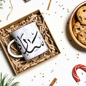 Puede incluir: Una taza de cerámica blanca con un diseño de caligrafía árabe negra, "سبحان الله", está anidada en una caja de regalo de papel marrón llena de papel triturado. La taza está rodeada de varios artículos festivos, incluyendo palitos de canela, bastones de caramelo y galletas con chispas de chocolate en un tazón de madera.