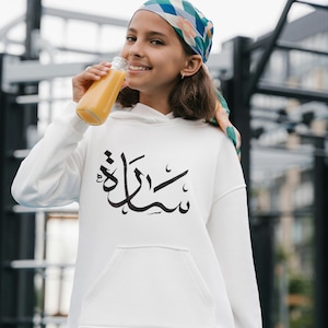 Könnte beinhalten: Eine junge Person trägt ein weißes Hoodie mit arabischer Kalligraphie, auf dem "Shukran" steht. Sie trägt einen rosa Rock und ein buntes Kopftuch. Sie hält eine Glasflasche Orangensaft in der Hand und lächelt.
